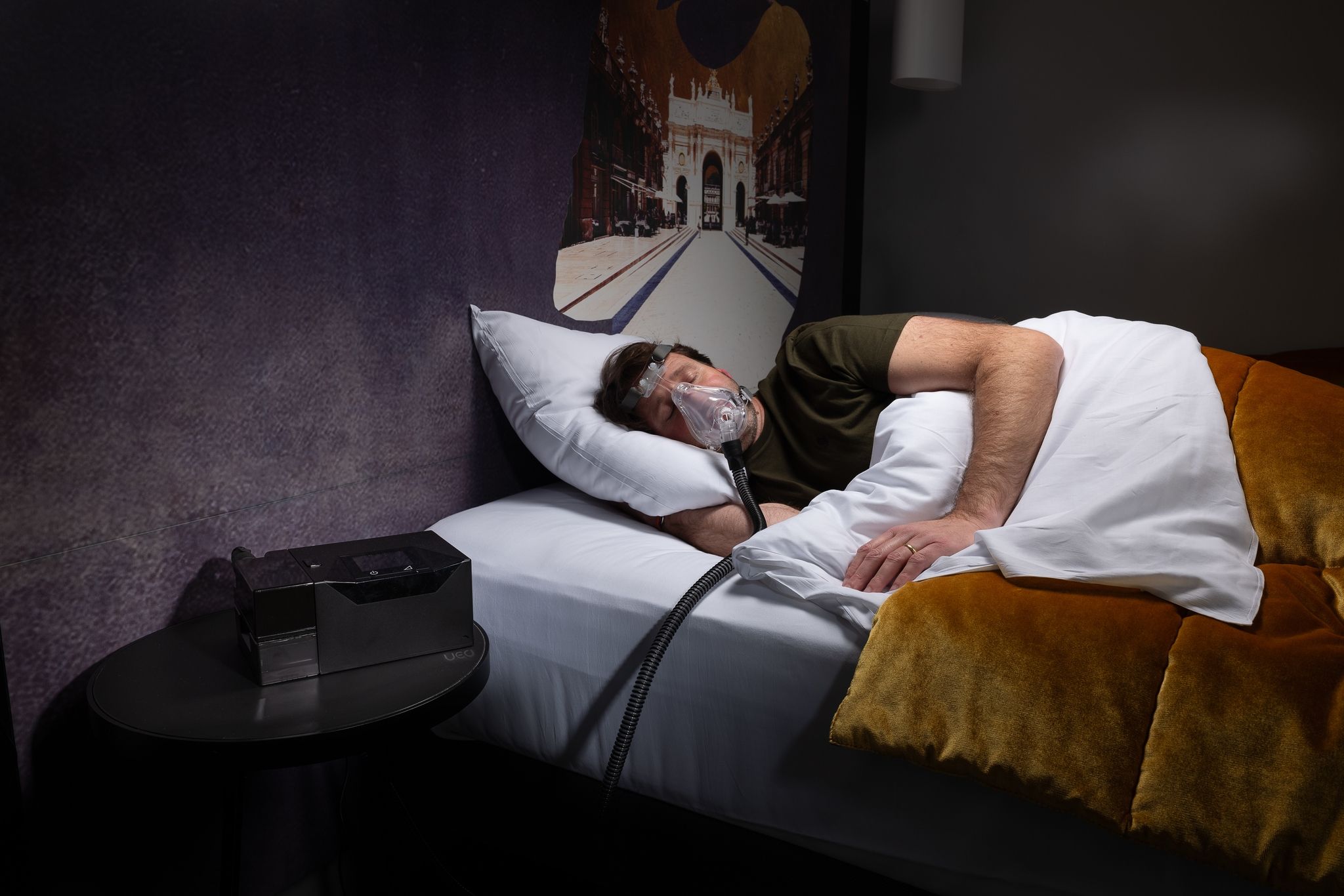Mann schläft mit Maske auf dem Bett. APAP-Gerät neben dem Bett.