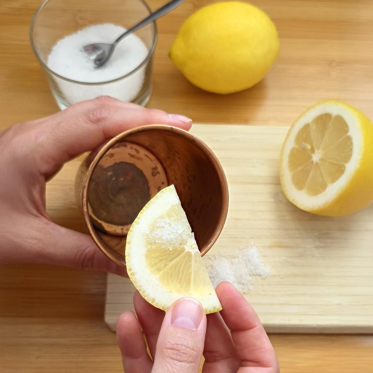 Zitrone und Kupferbecher. Zitrone wird in Becher gegeben. Holzbrett und Zucker im Hintergrund.