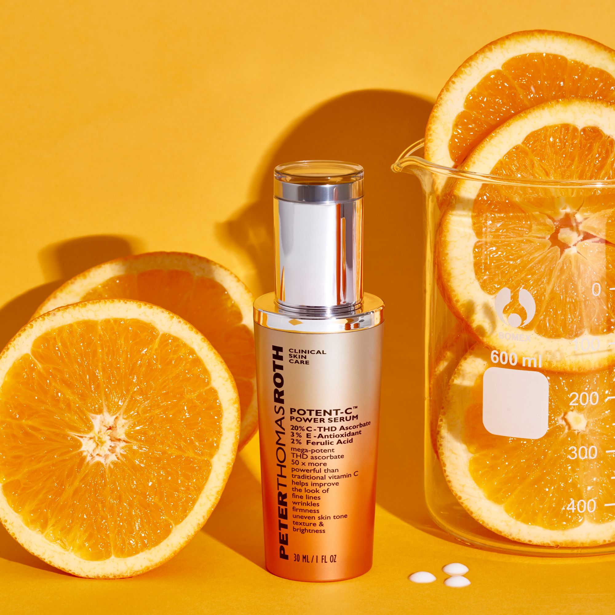 Serumflasche mit Orangen und Becherglas. Aufschrift: Potent-C Power Serum. Silberne Kappe und orangefarbener Farbverlauf. Orangenstücke.
