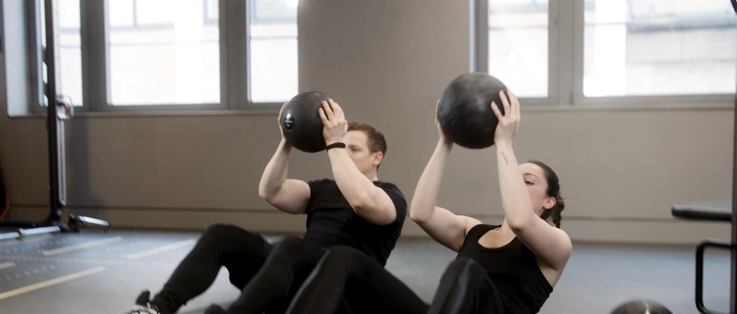 Zwei Personen sitzen und heben jeweils einen schwarzen Slamball. Sie befinden sich in einem Fitnessstudio.
