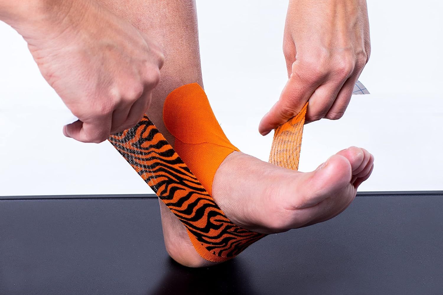 Hände applizieren CureTape Tiger am Fuß. Orange mit schwarzen Streifen. Auf schwarzem Untergrund. Tape wird angebracht.