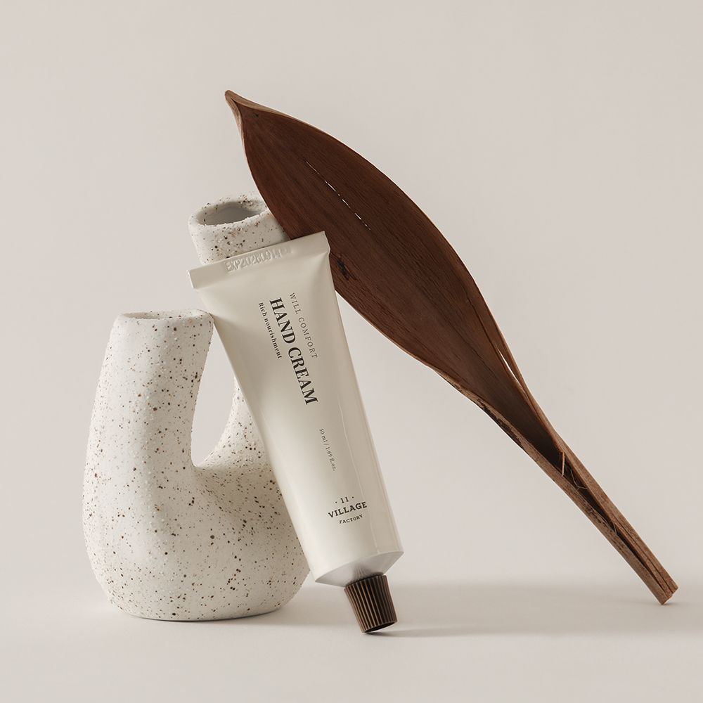 Creme-Tube, neben einer Vase und einem Blatt. Aufschrift: HAND CREAM, VILLAGE FACTORY. Beige Tube mit braunem Verschluss.