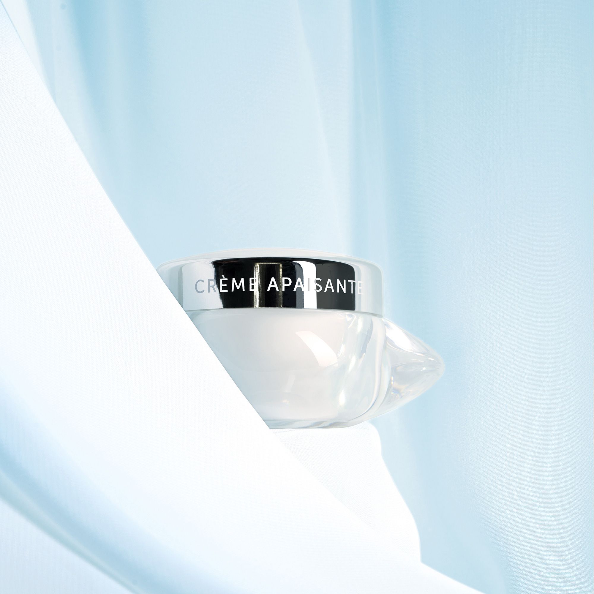Glasbehälter mit silbernem Deckel. Aufschrift: CRÈME APAISANTE. Vor blauem Stoff platziert.