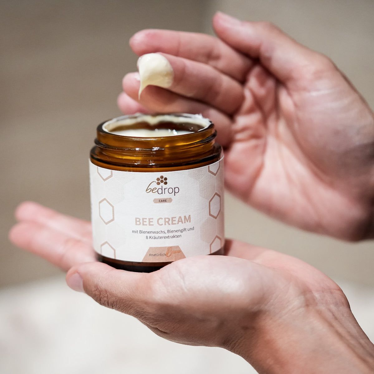 Hände halten braune Glasdose "bedrop Bee Cream". Creme auf Finger. Hintergrund unscharf.