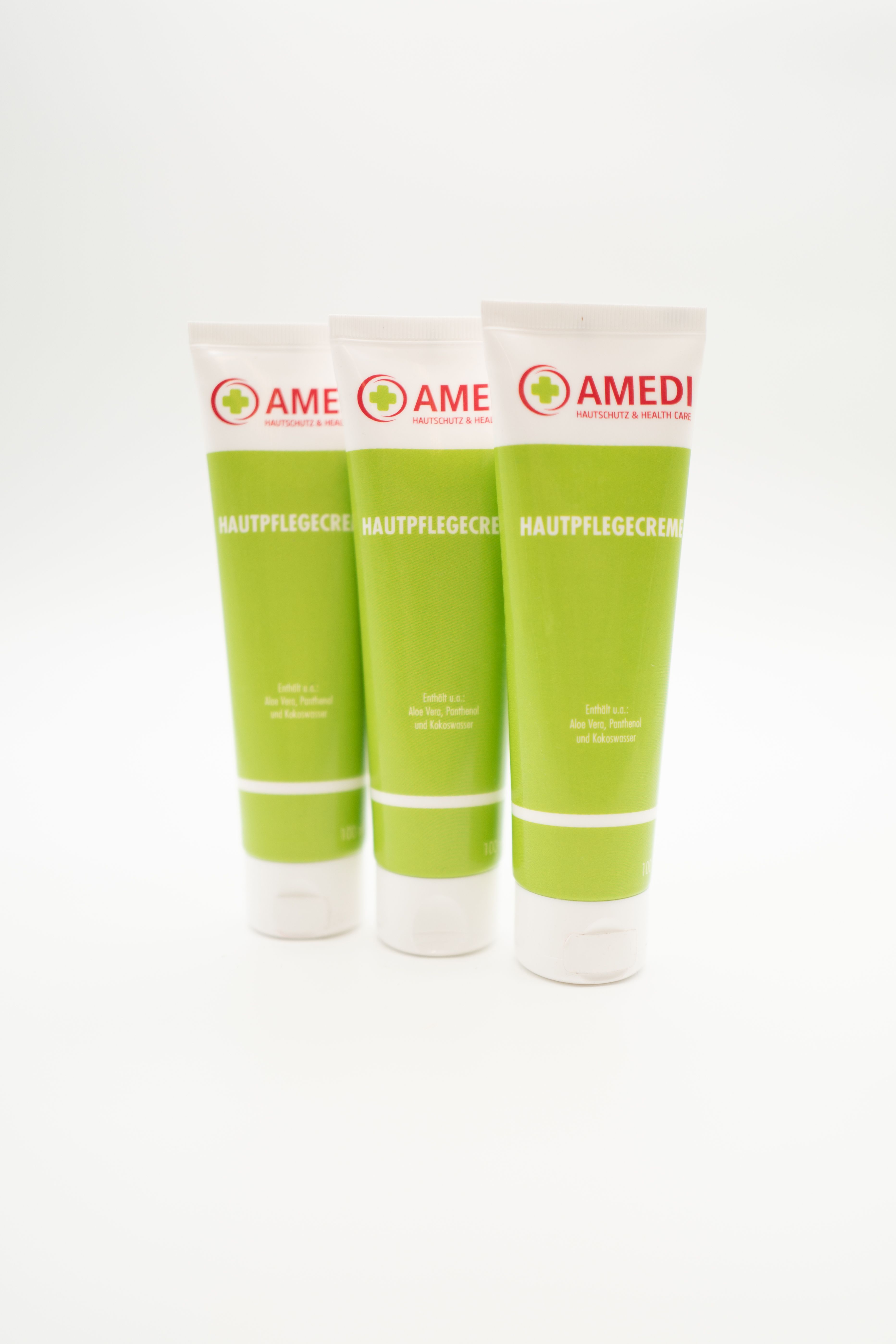 Drei grüne Tuben mit weißem Deckel. Aufschrift: AMEDI, Hautpflegecreme. Logo mit rotem Kreuz. Text in Deutsch.