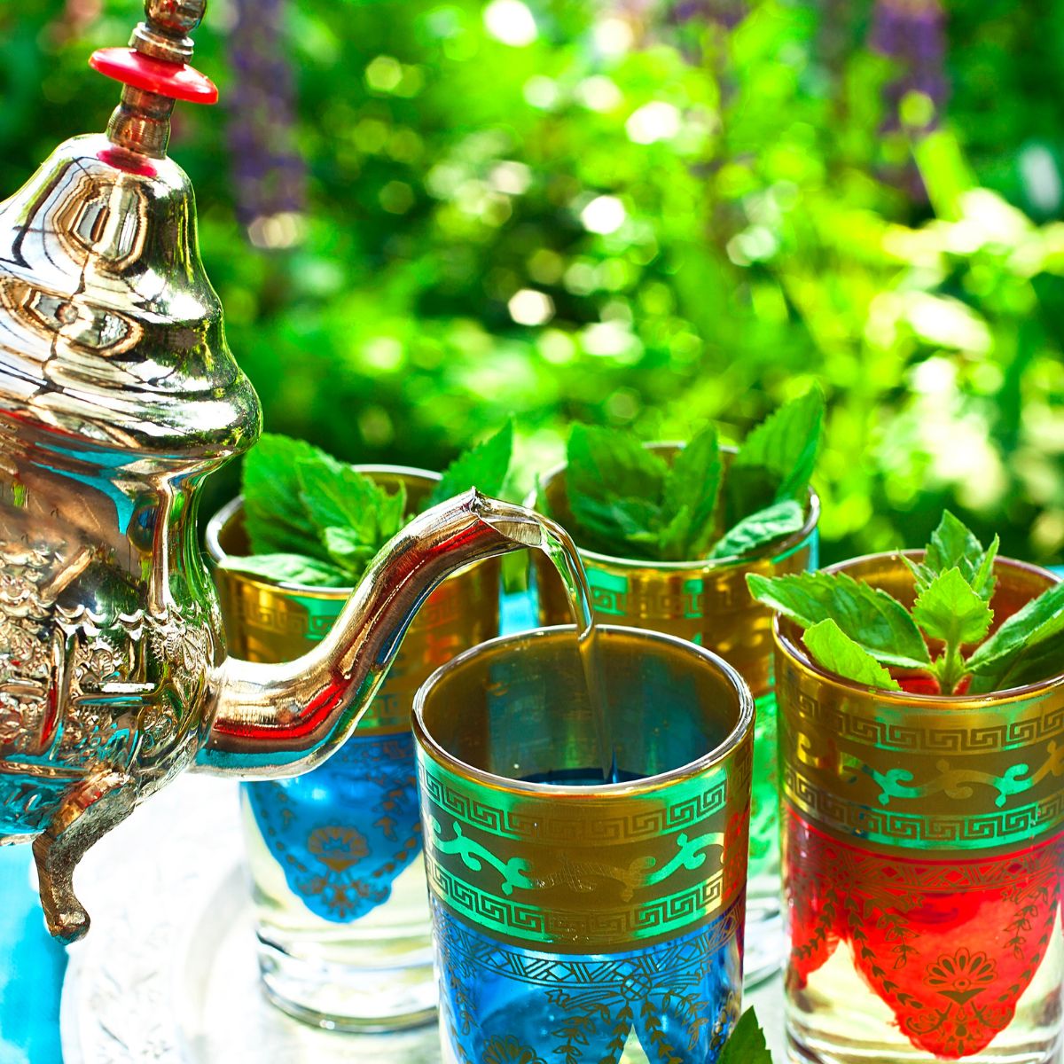 Teekanne gießt Tee in Gläser mit Minzblättern. Gold- und blau verzierte Gläser. Grüner Hintergrund.