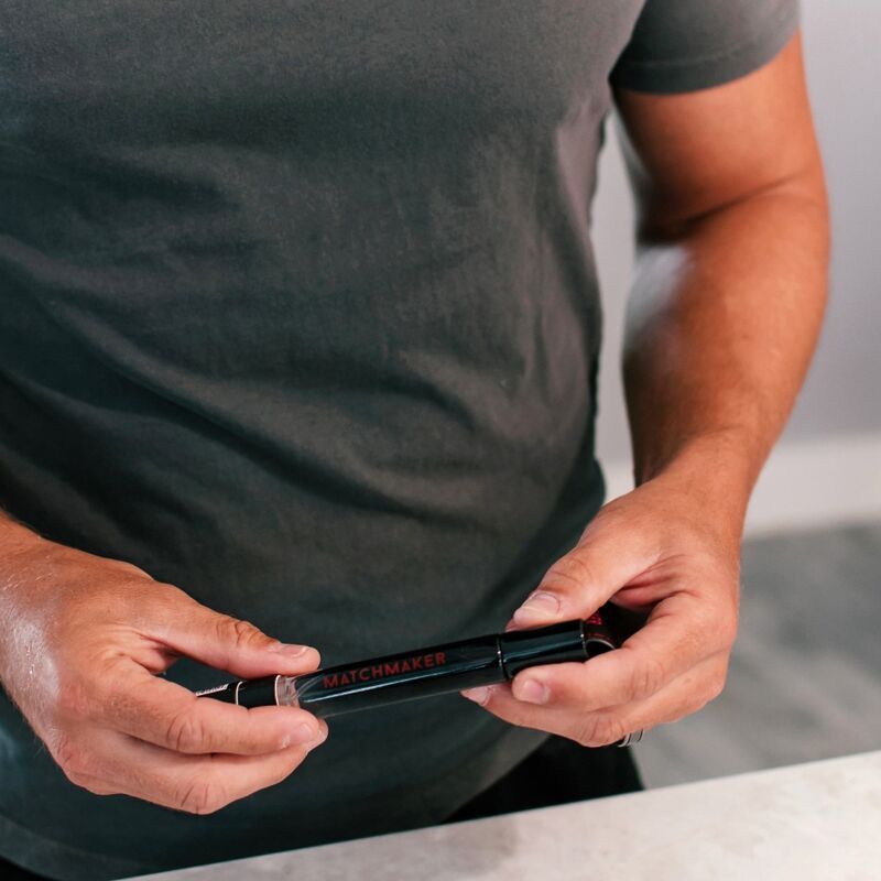Mann hält einen schwarzen Parfümflakon. Rote Schrift "MATCHMAKER" auf dem Flakon. Der Mann trägt ein graues T-Shirt.