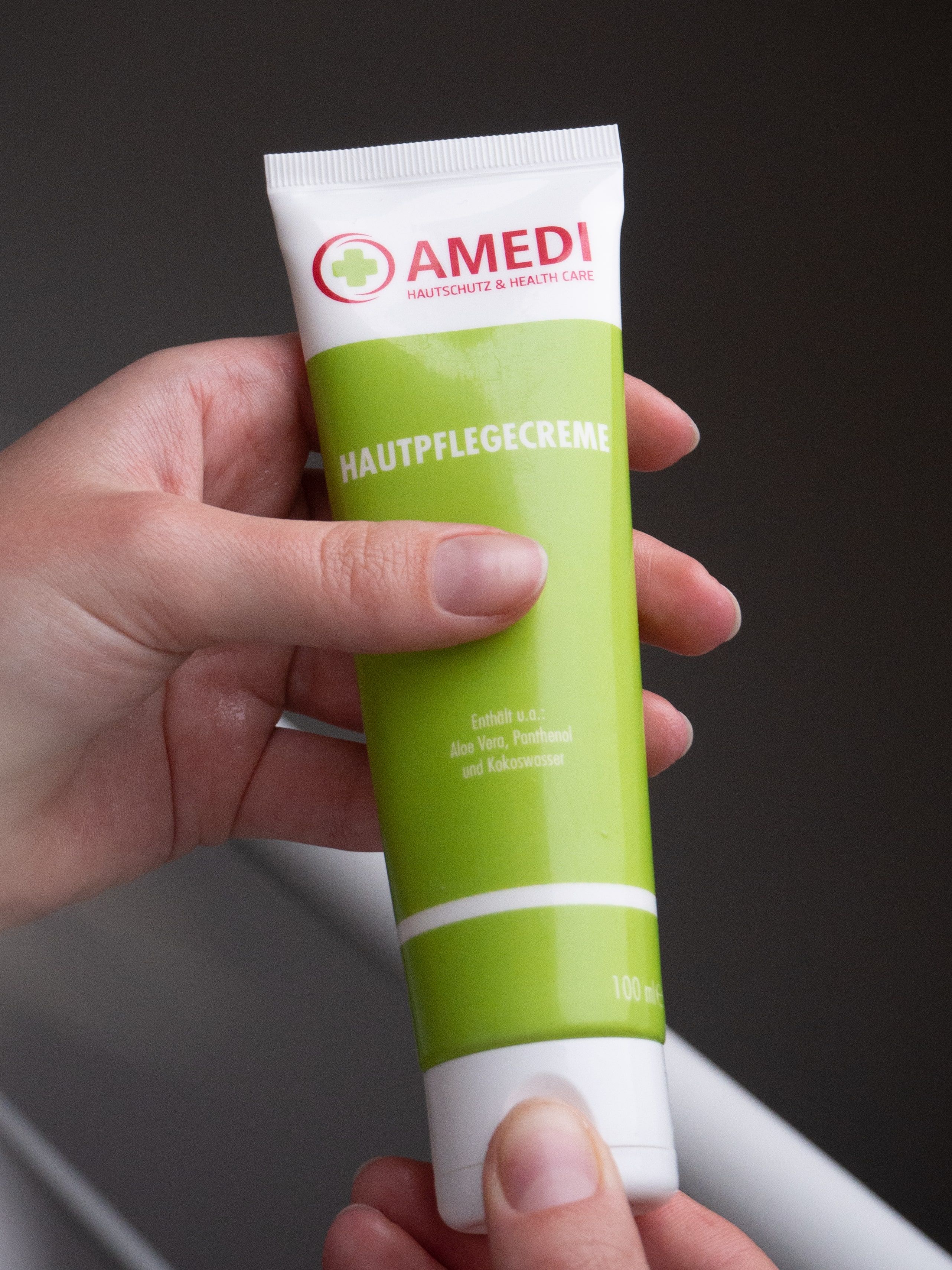 Hand hält grüne Tube mit weißem Deckel. Aufschrift: AMEDI, Hautpflegecreme. Logo mit rotem Kreuz. Text in Deutsch.