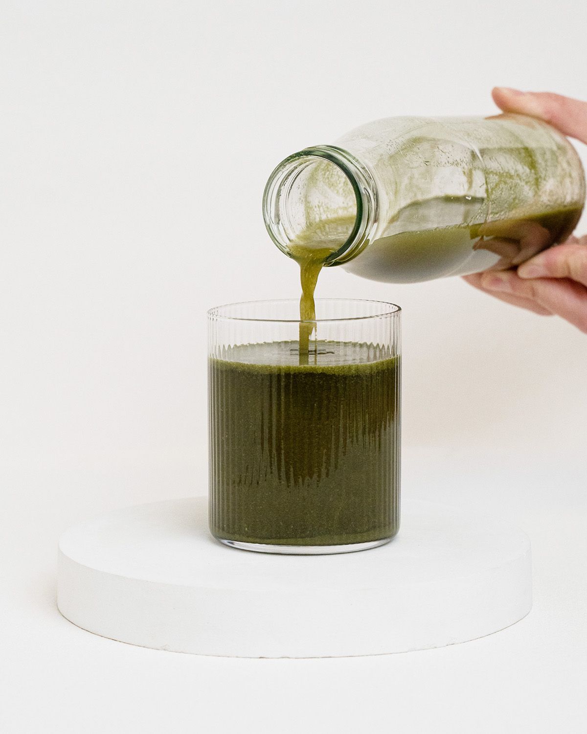 Grüne Flüssigkeit wird aus einer Glasflasche in ein geriffeltes Glas gegossen. Auf weißem Podest.