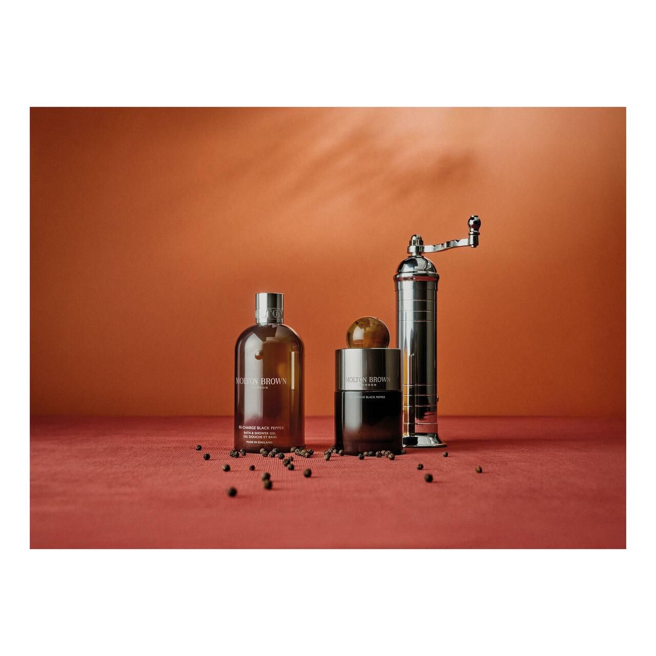 Produktflasche, Tiegel und Pfeffermühle. Schwarze Pfefferkörner auf rotem Untergrund. Marke Molton Brown.