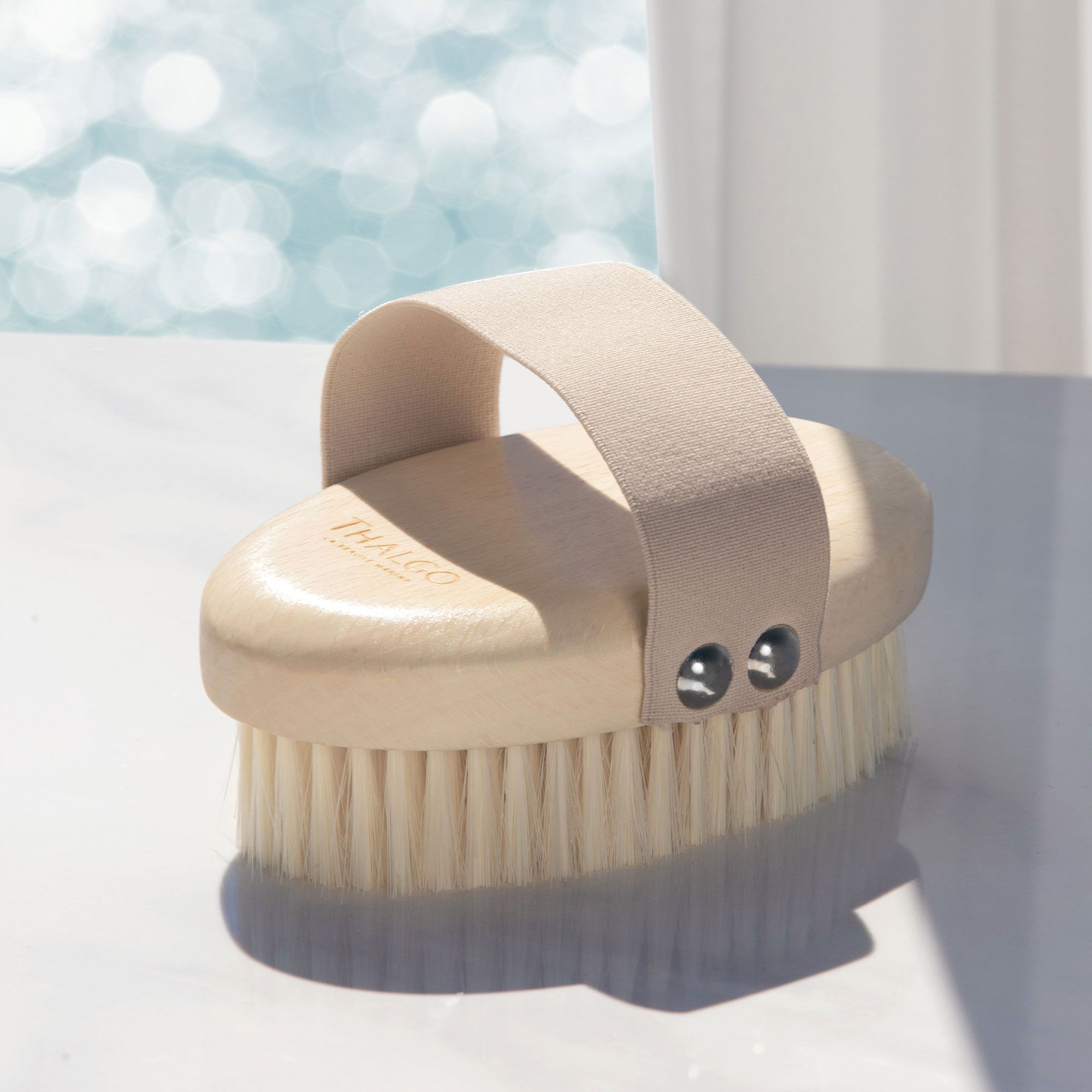 THALGO Massagebürste. Ovalförmige Bürste mit hellem Holzgriff, hellen Borsten und Lederband. Auf weißer Oberfläche.
