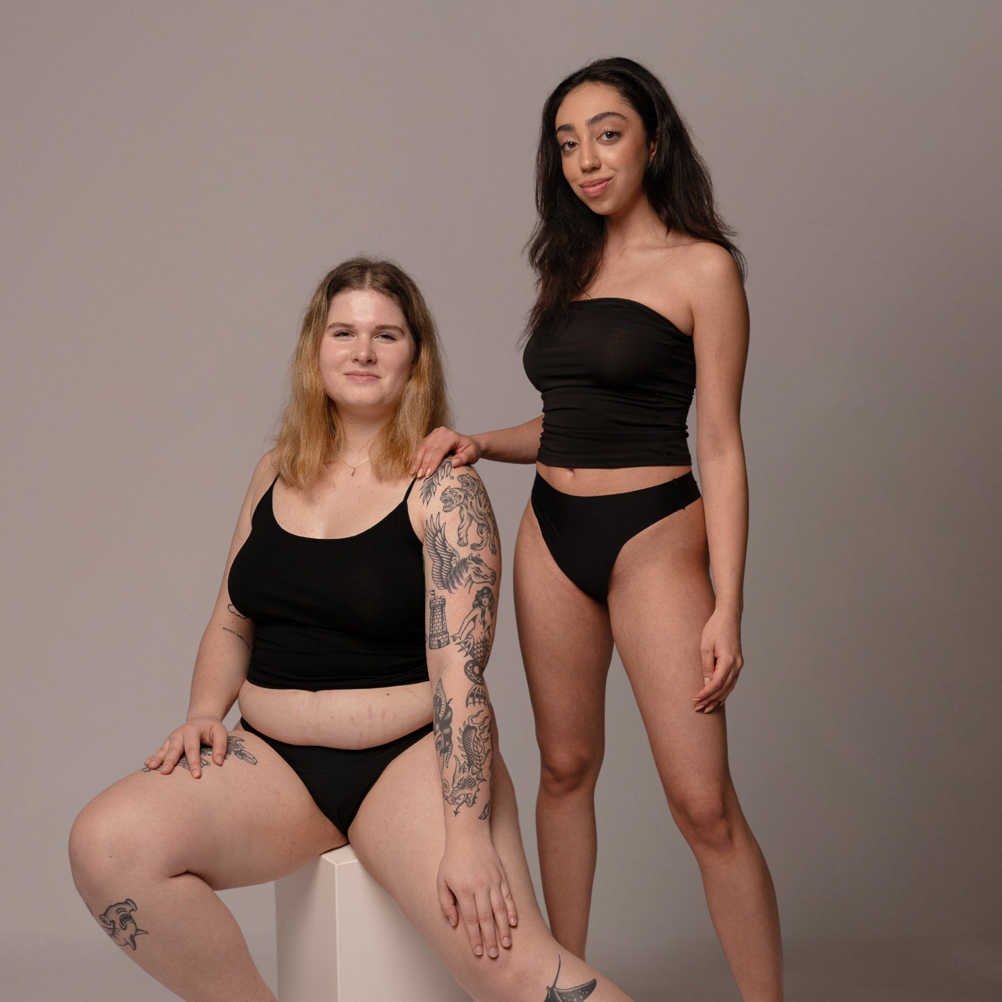 Zwei Frauen in schwarzen Tanktops und Slips. Eine sitzt, die andere steht.