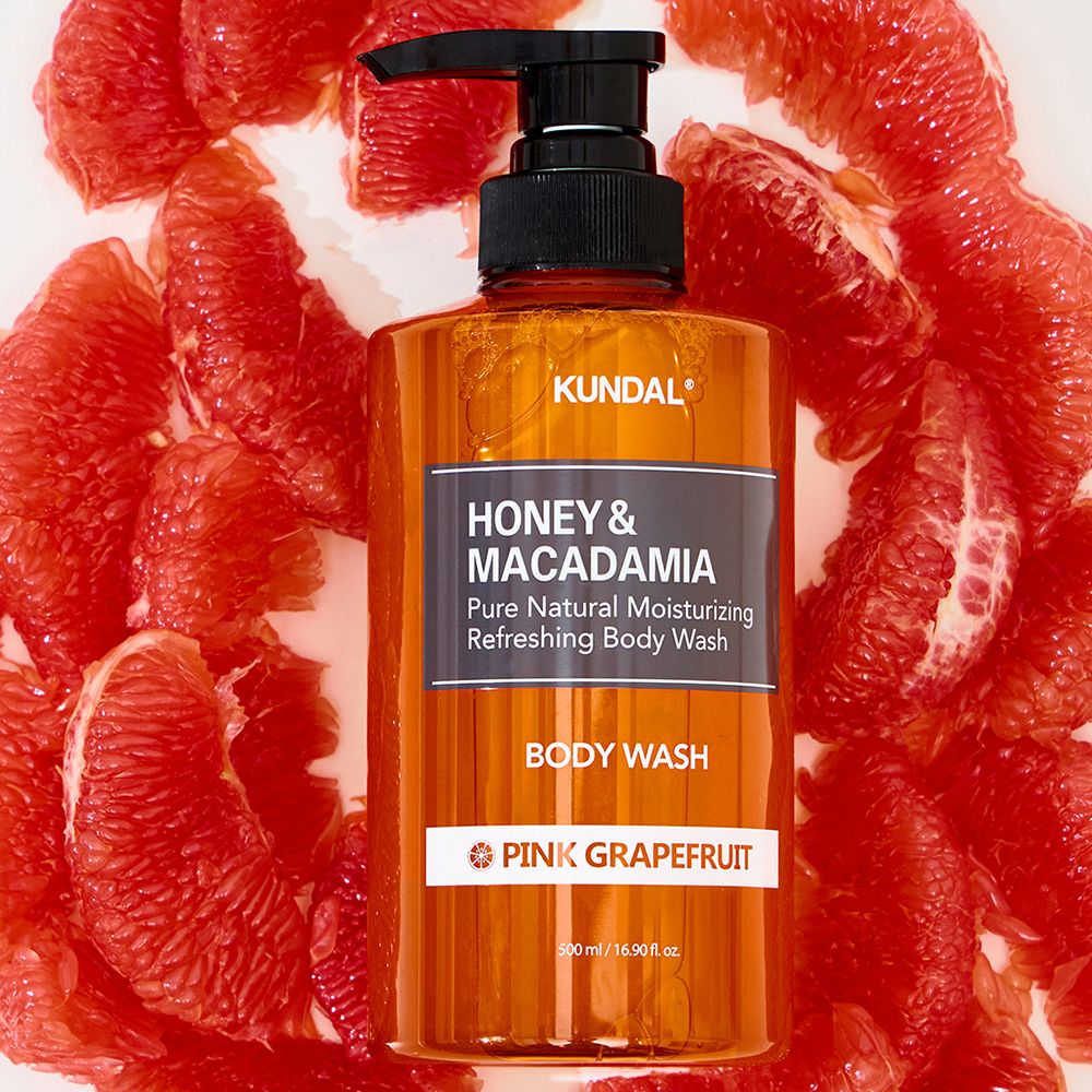 Orangefarbene Flasche mit schwarzem Pumpverschluss, umgeben von rosa Grapefruit-Stücken. Aufschrift: KUNDAL, HONEY & MACADAMIA.
