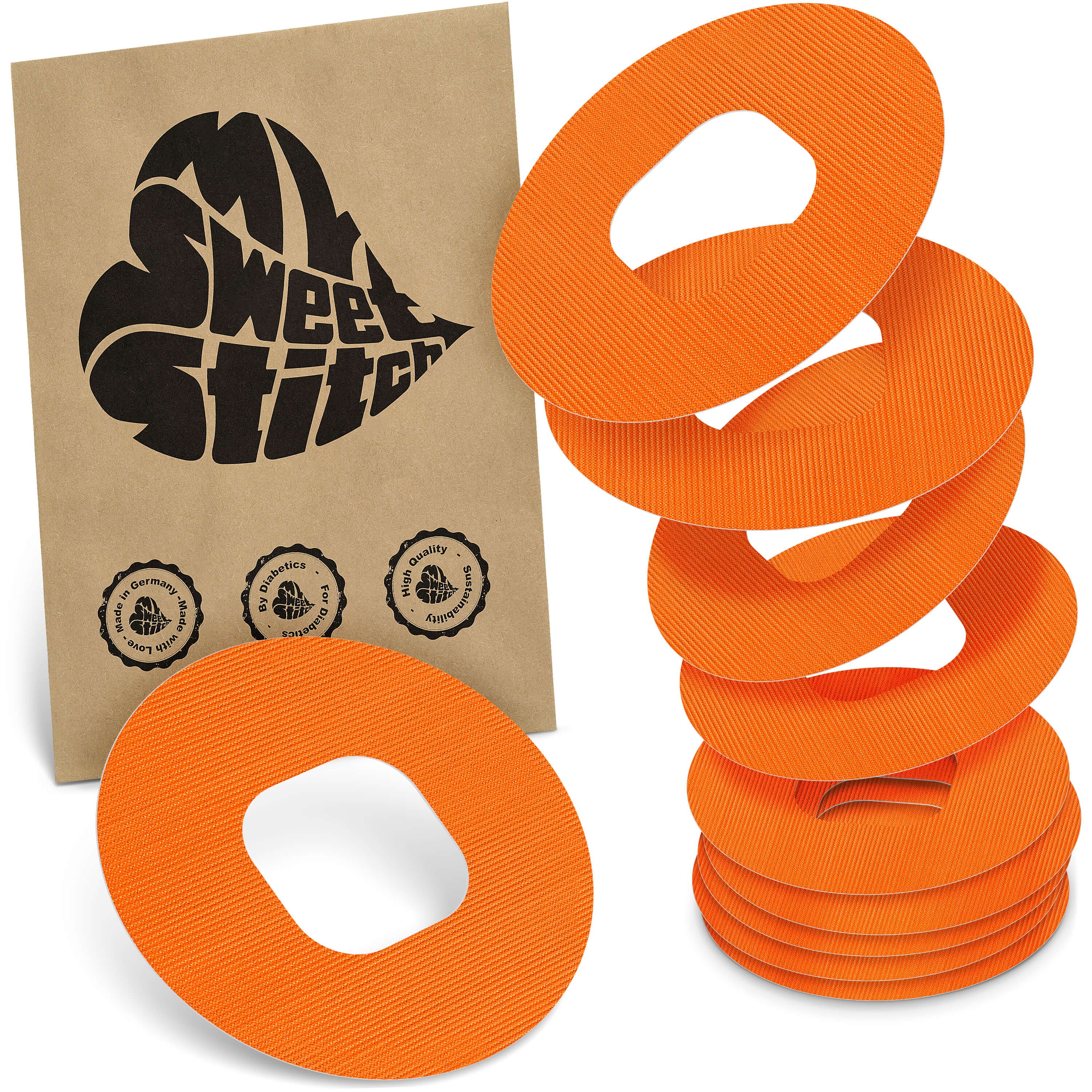 Orange, ringförmige Pflaster mit einer Aussparung in der Mitte. Ein Stapel von sechs Pflastern liegt rechts, ein einzelnes liegt links. Daneben eine braune Papiertüte mit schwarzem Herz-Logo.