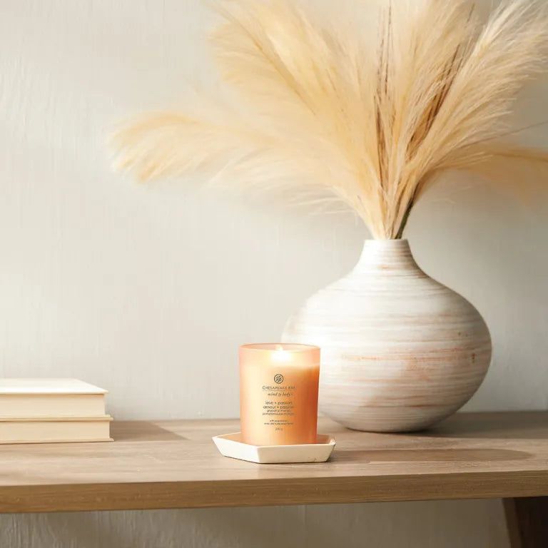 Kerze in Glasbehälter auf weißem Untersetzer. Neben Vase mit Pampasgras. Auf Holzoberfläche.