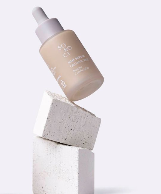 Serumflasche auf Steinblöcken. Weißer Deckel, Aufschrift. Beige Flasche. Aufschrift: NMF Serum Organic Rice.