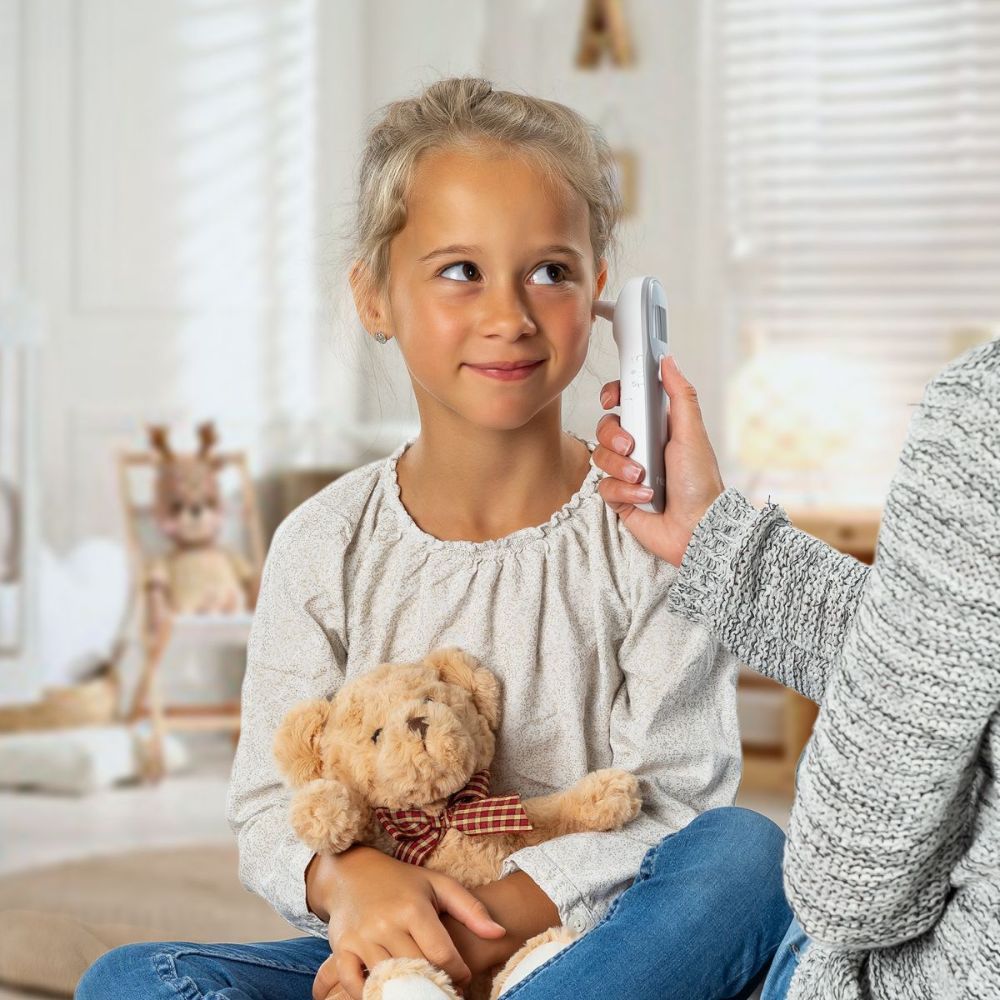 Kind mit Ohrthermometer am Ohr. Person hält Thermometer. Teddybär im Hintergrund.