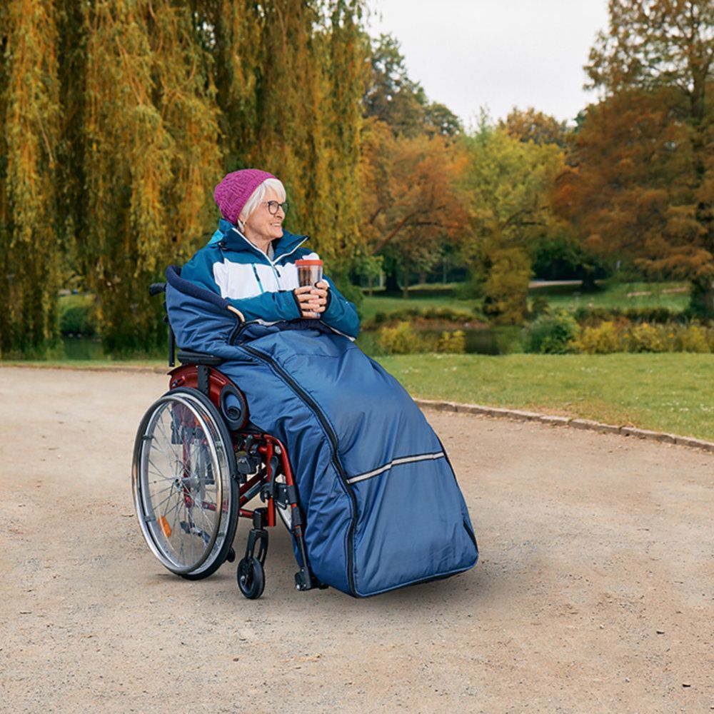 Blauer Schlupfsack für Rollstuhl. Ältere Frau im Rollstuhl im Park. Sie hält einen Becher. Herbstliche Umgebung.