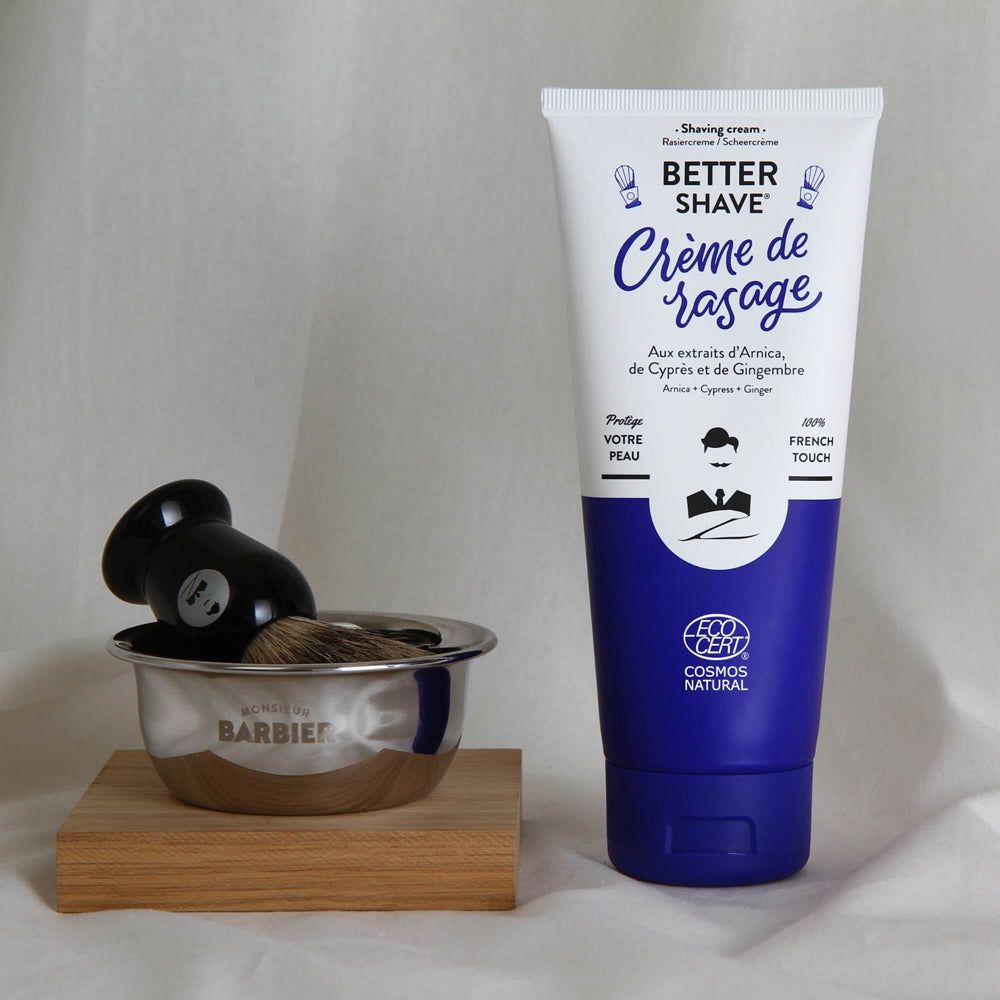 Rasiercreme-Tube mit Rasierpinsel und Schale auf Holz. Tube mit blauer Basis, weißem Hintergrund und Text. Schale mit BARBIER-Logo.