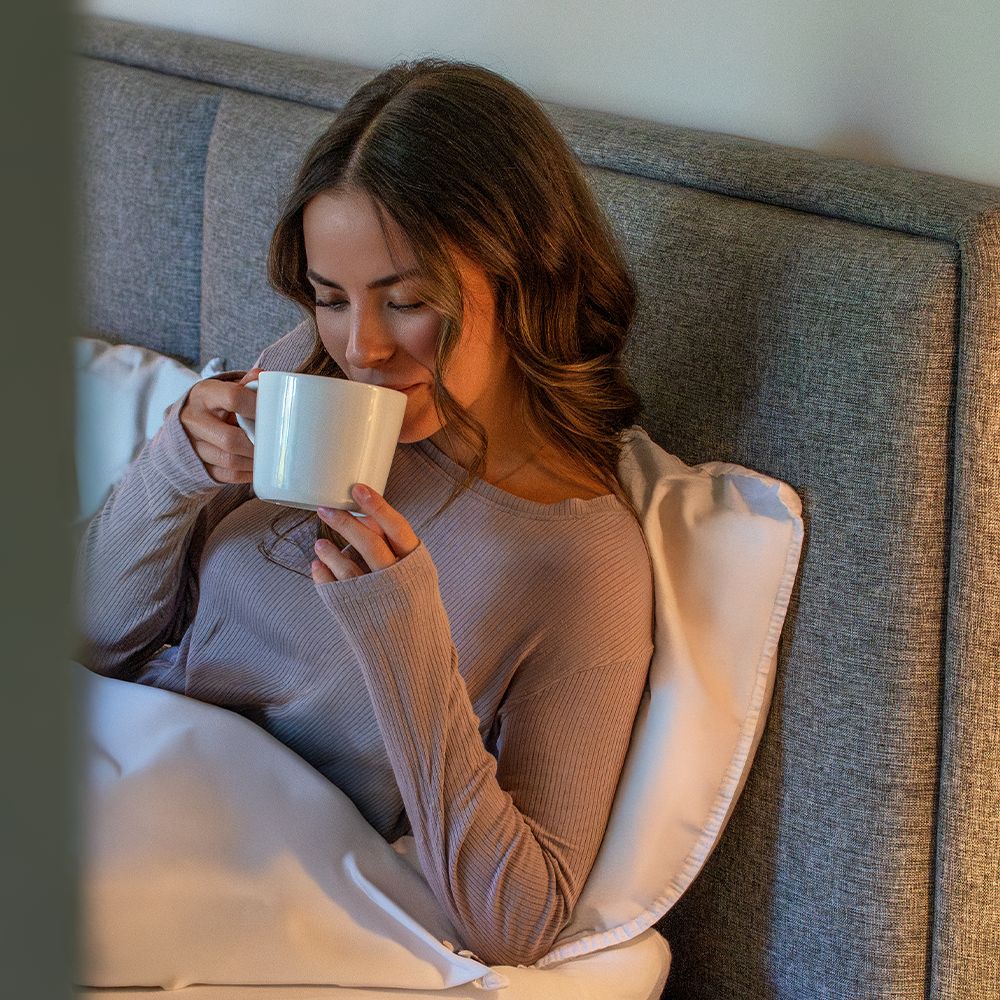 Person im Bett trinkt aus einer weißen Tasse. Im Hintergrund eine graue Wand.