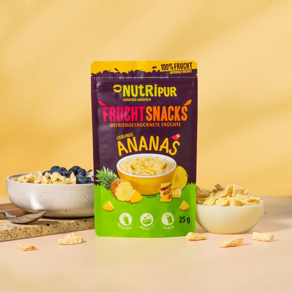 Beutel mit gefriergetrockneten Ananas-Bits. Daneben eine Schale mit Müsli und Blaubeeren. Aufschrift: NutriPur, Fruchtsnacks.