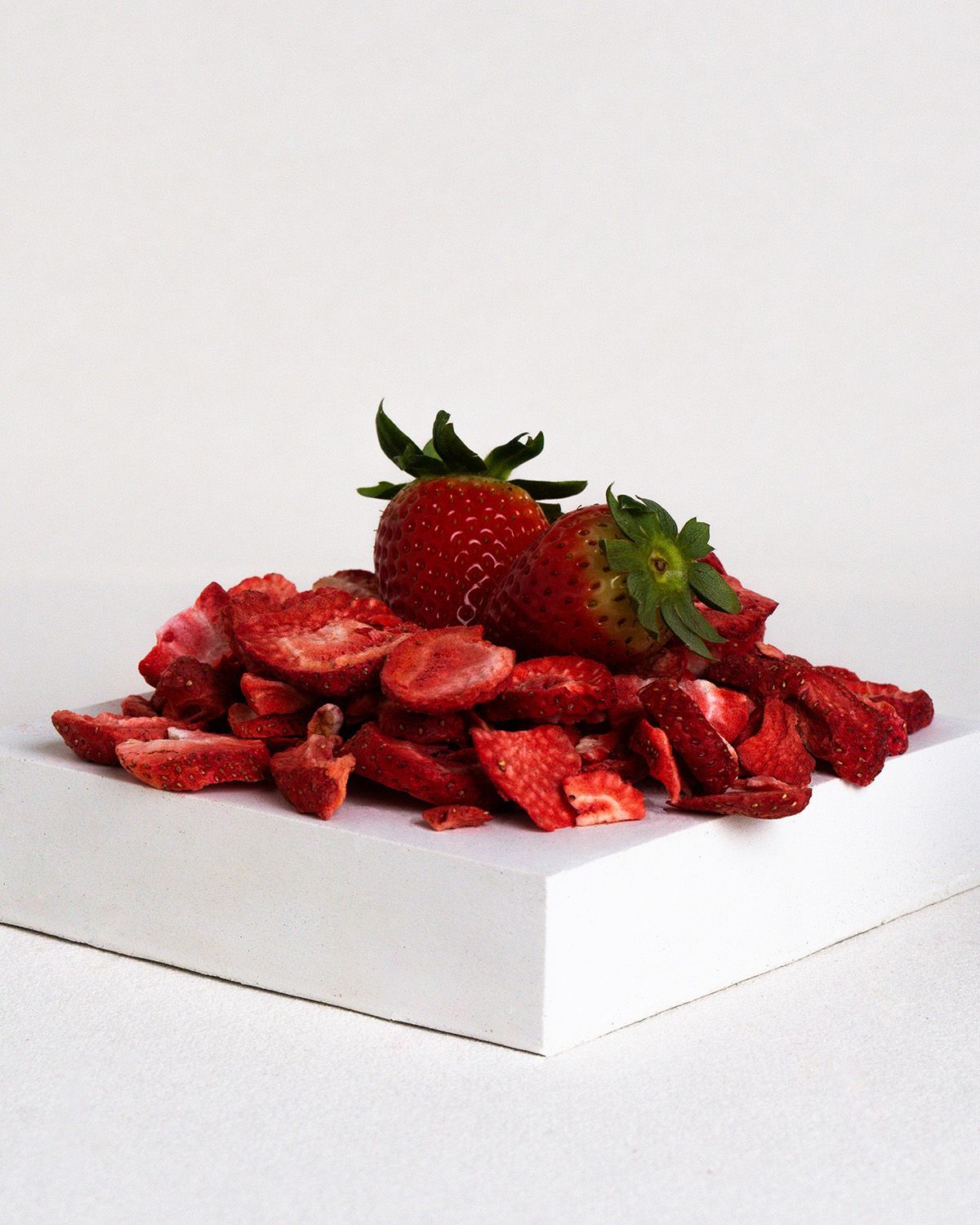 Haufen gefriergetrockneter Erdbeerscheiben auf weißem Podest. Obenauf zwei frische Erdbeeren mit grünen Blättern.