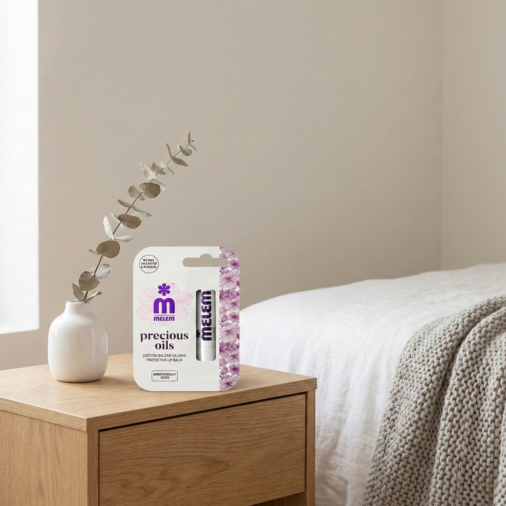 Melem Schützender Lippenbalsam in Verpackung. Weißes Produkt mit lila Akzenten. Auf Nachttisch neben Vase und Bett.