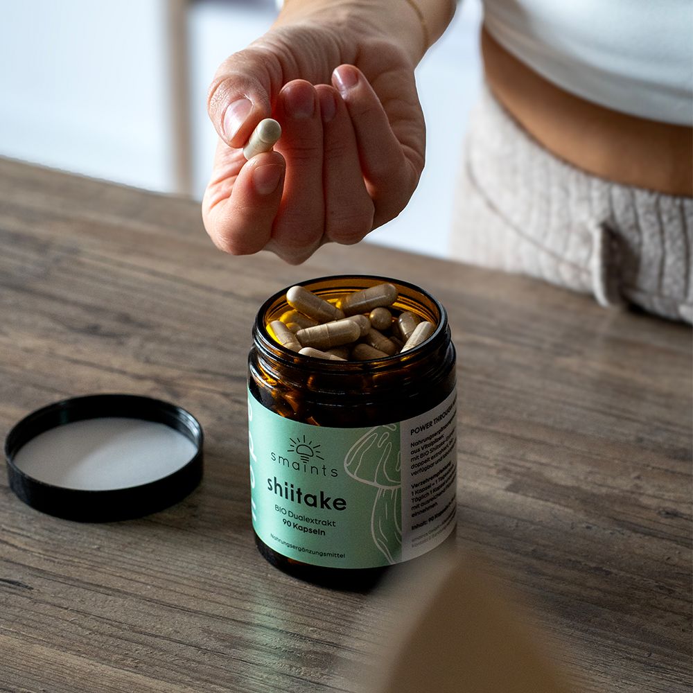 Offene braune Glasflasche mit Kapseln. Eine Hand hält eine Kapsel. Auf dem Tisch liegt der Deckel.
