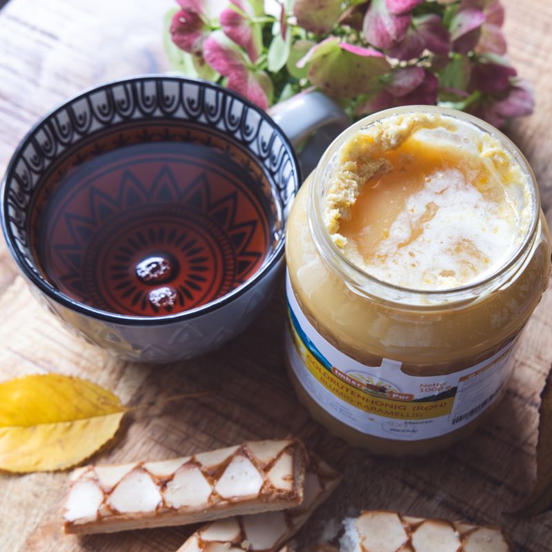 Glas Honig mit Etikett, Goldrutenhonig (roh), neben Tasse Tee und Gebäck. Deckel goldfarben.