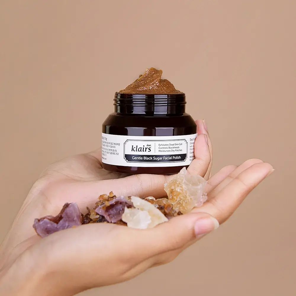 Glas mit brauner Creme, gehalten von Händen. Daneben Steine. Aufschrift: Klairs Gentle Black Sugar Facial Polish.