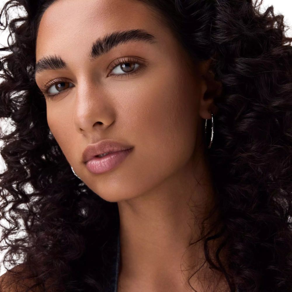 Porträt einer Frau mit dunklen Locken und silbernen Ohrringen. Hautton und Augenfarbe sind sichtbar. Keine Produkte sichtbar.