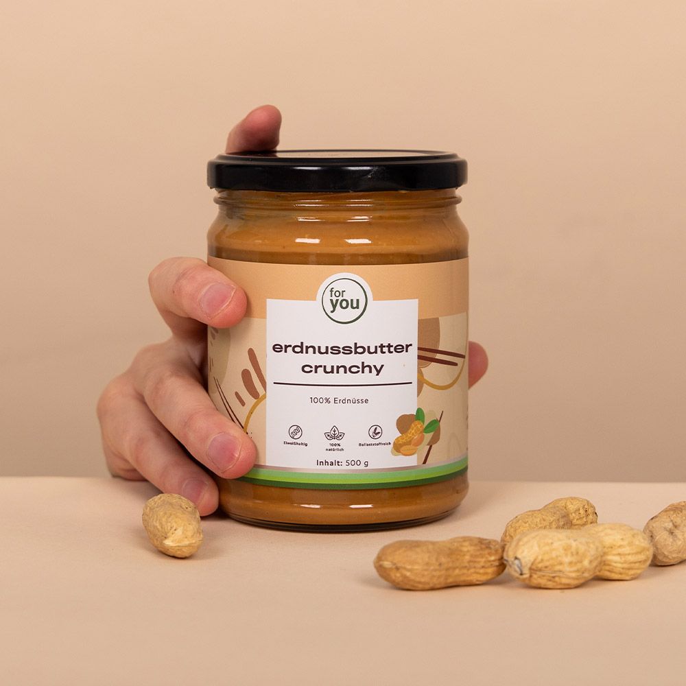 Glas Erdnussbutter Crunchy in Hand. Aufschrift: for you, Erdnussbutter Crunchy, 100% Erdnüsse, Inhalt: 500 g. Daneben Erdnüsse.