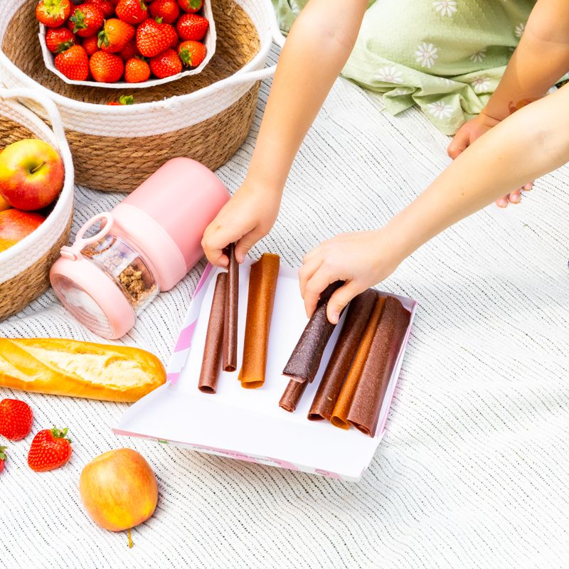 BIO Fruchtrollen und Früchte auf Picknickdecke. Kind greift nach Rollen. Weitere Lebensmittel sichtbar.