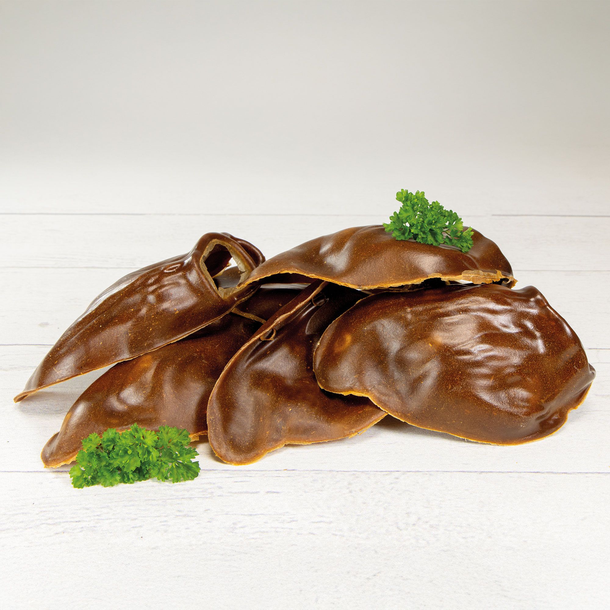 Mehrere braune, ohrförmige Kauartikel, mit Petersilie garniert, auf weißem Holzgrund.