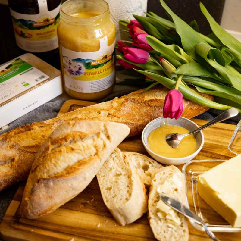 Bergblütenhonig-Glas, Brot, Butter, Honig in Schale, Blumen. Holzbrett.