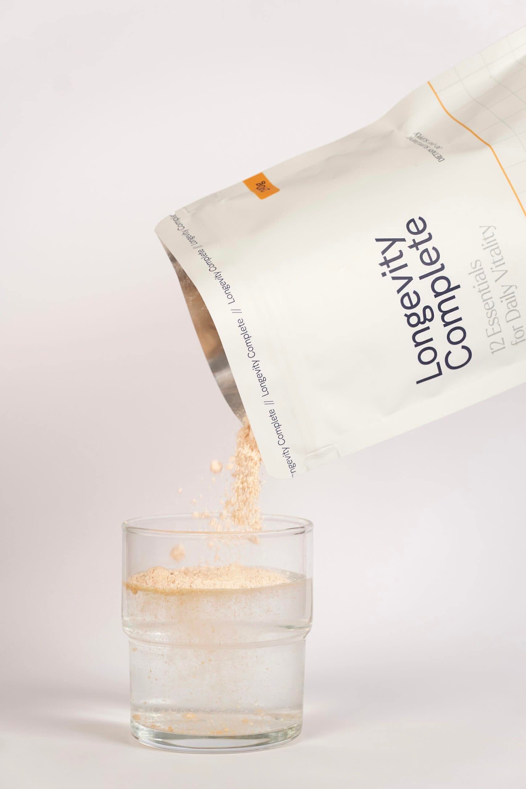 Ein Beutel mit Pulver wird über ein Glas Wasser gegossen. Der Beutel ist mit 'Longevity Complete' und '12 Essentials for Daily Vitality' beschriftet.