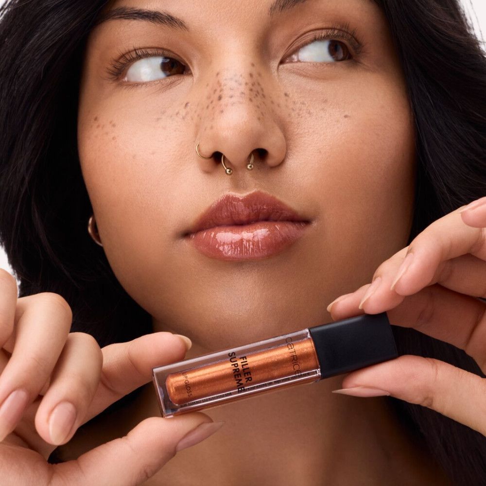 Frau hält Lipgloss-Tube. Aufschrift: CATRICE, FILLER SUPREME. Lippen mit Lipgloss. Bronzefarbener Lipgloss.