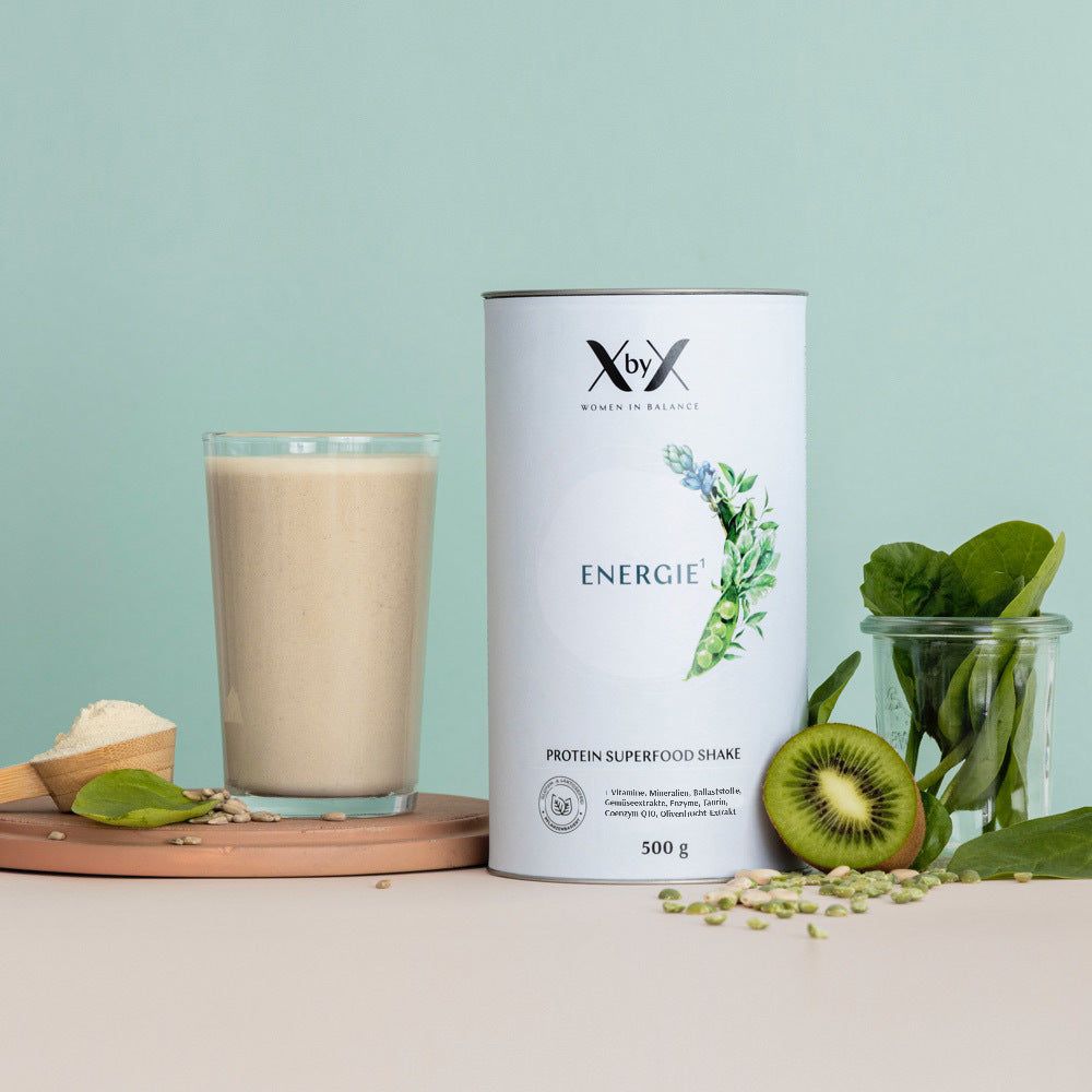 Weißes Produkt-Behälter mit Aufschrift 'XbyX Energie'. Daneben Glas mit Getränk, Holzlöffel mit Pulver, Kiwi, Spinat.