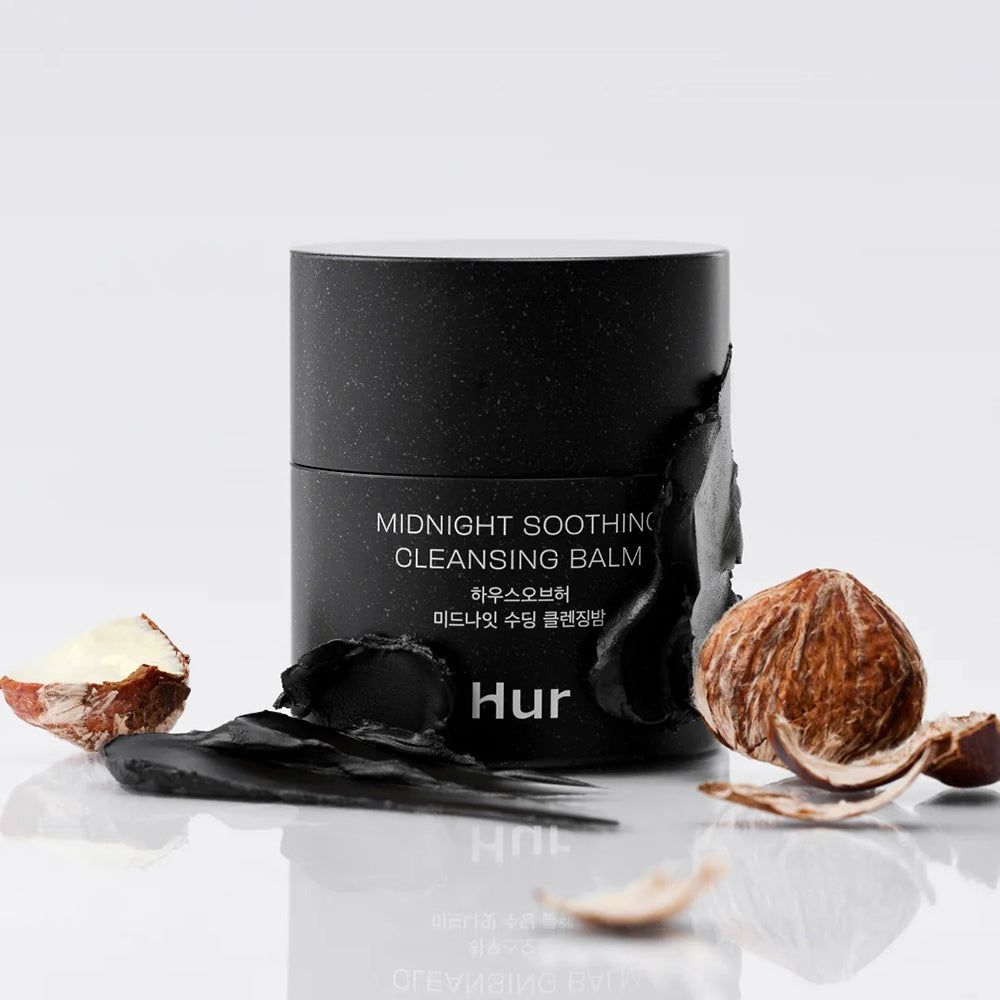 Schwarzer Tiegel mit Aufschrift: Midnight Soothing Cleansing Balm. Marke: Hur. Umgeben von Zutaten. Hintergrund weiß.