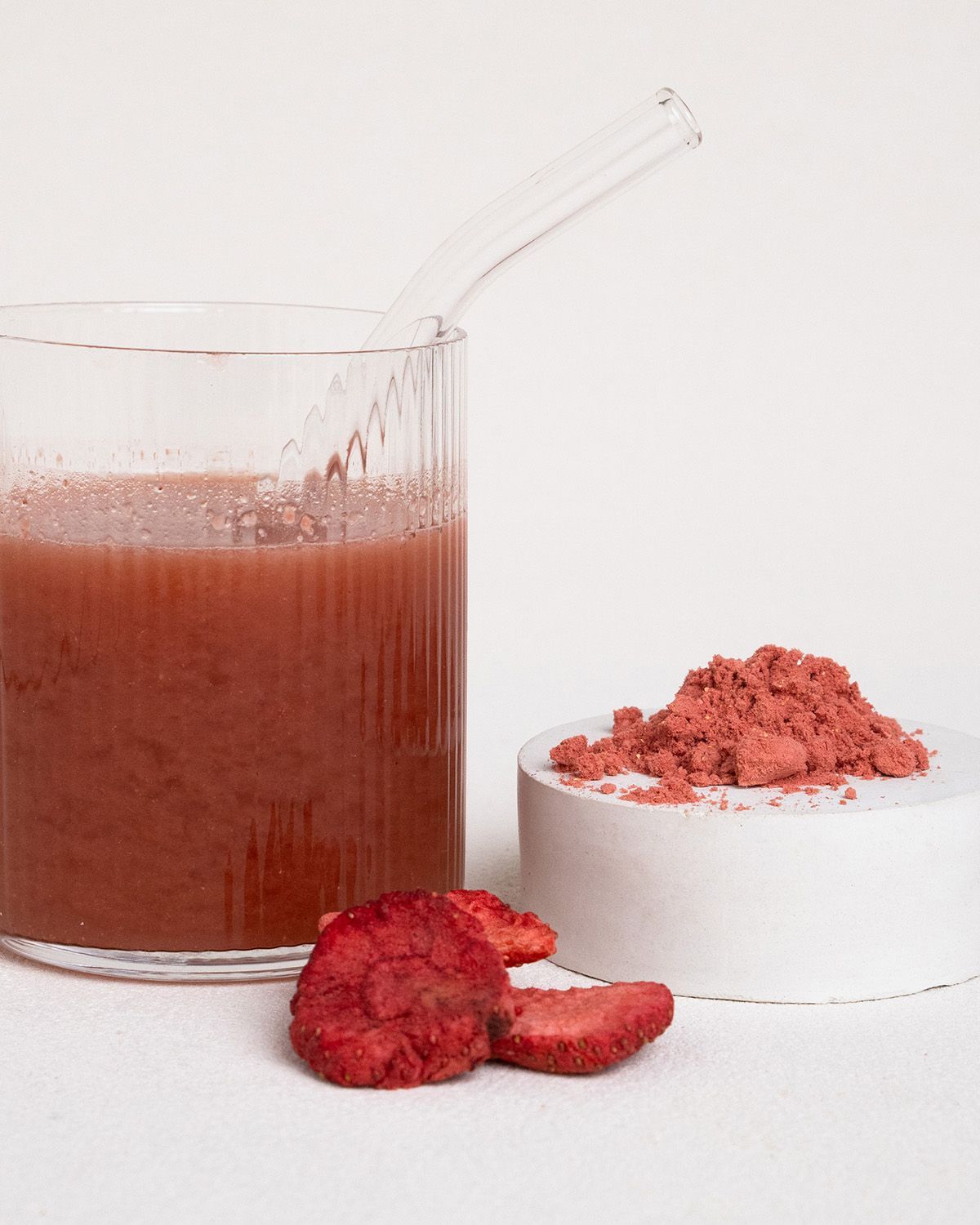 Glas mit Erdbeergetränk, Erdbeerpulver auf einem Podest und getrocknete Erdbeeren.