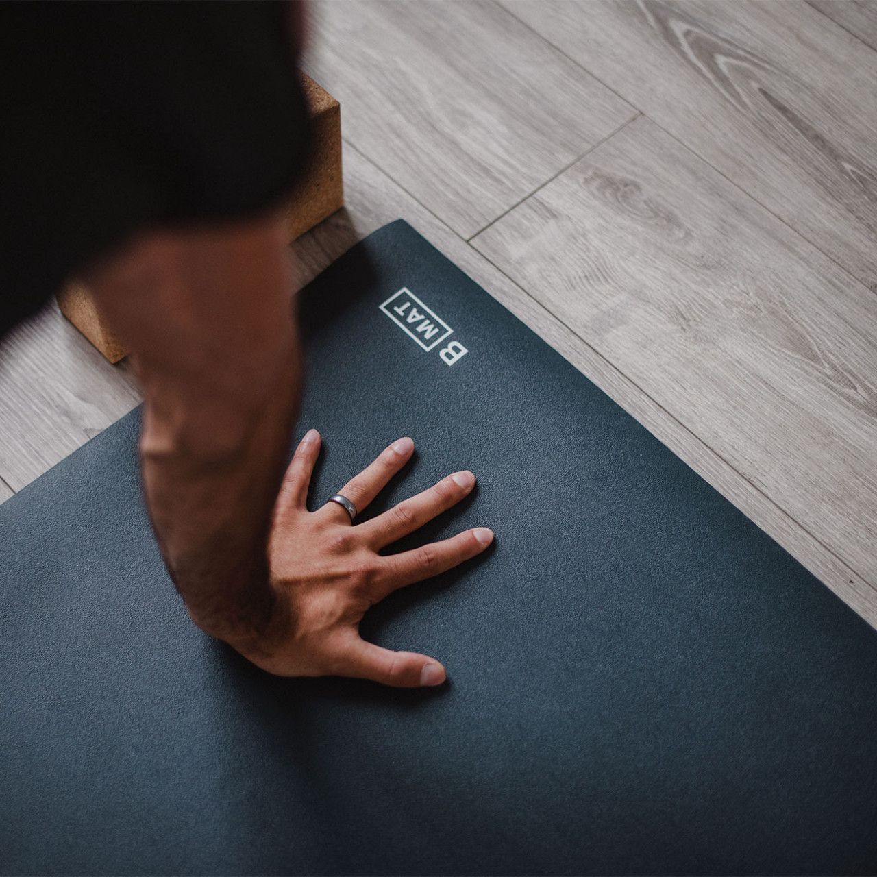Hand auf grauer Yogamatte mit Logo "B MAT". Holzblock im Hintergrund.