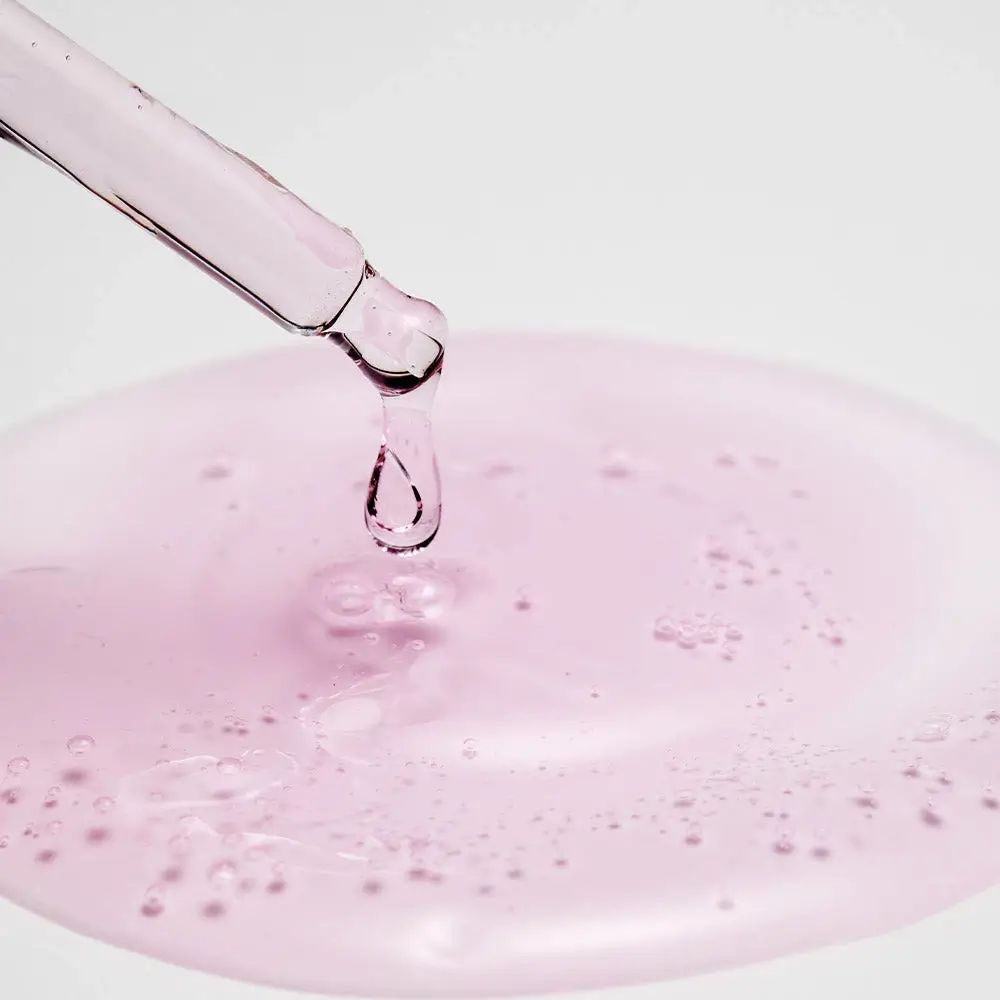 Pipette mit rosa Flüssigkeit, die in eine Pfütze tropft. Flüssigkeit mit Bläschen. Hintergrund weiß.