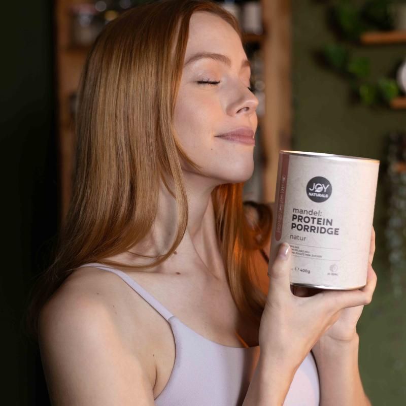 Frau hält Dose 'JOY NATURALS Mandel Protein Porridge Natur'. Dose mit Logo und Text. Frau mit geschlossenen Augen.