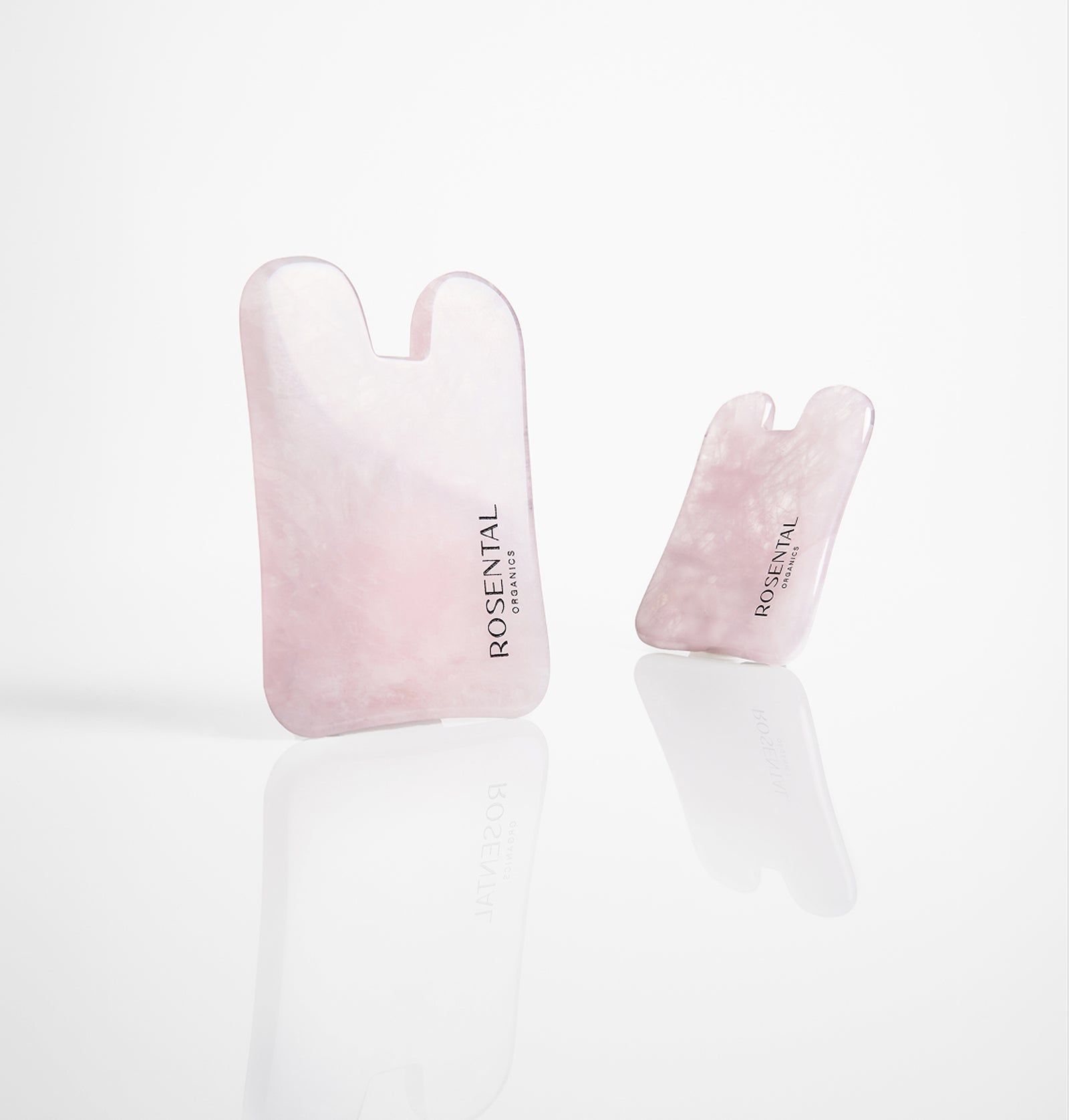 Zwei Gua Shas aus Rosenquarz. Rosa Steine, abgerundete Form mit zwei Aussparungen. Text: Rosental Organics. Reflektierender Untergrund.