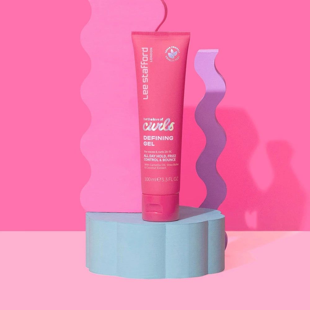 Rosa Tube Lee Stafford For The Love Of Curls Defining Gel auf einem blauen Podest vor rosa Hintergrund.