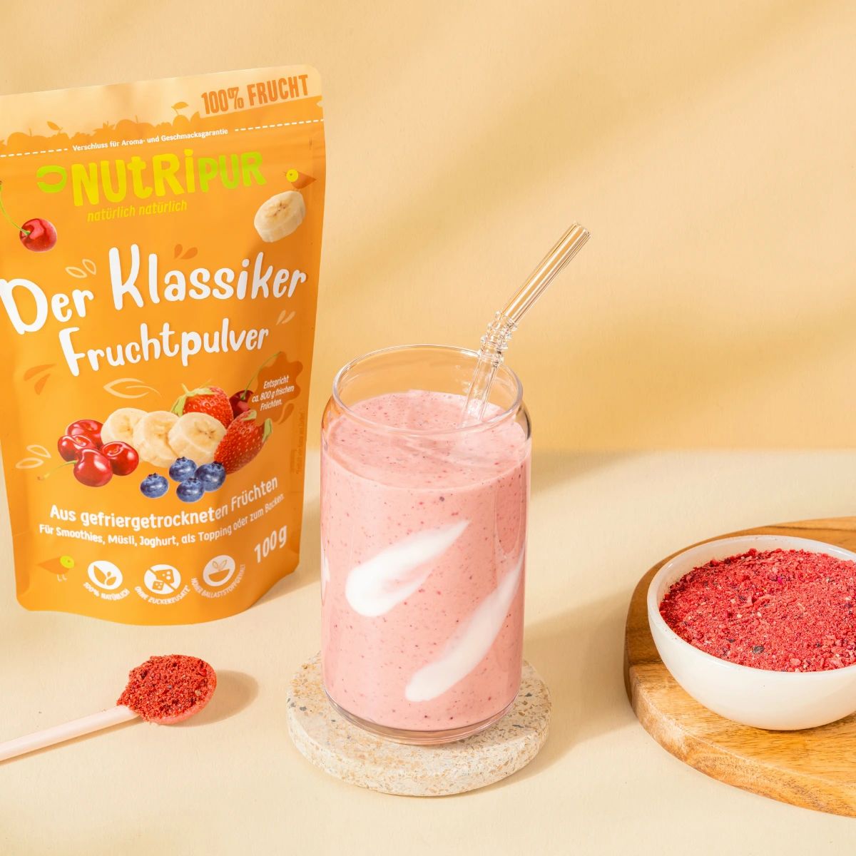 Glas mit rosa Smoothie, Strohhalm, daneben Verpackung NutriPur Fruchtpulver und Schale mit rotem Pulver. Auf einem Untersetzer.
