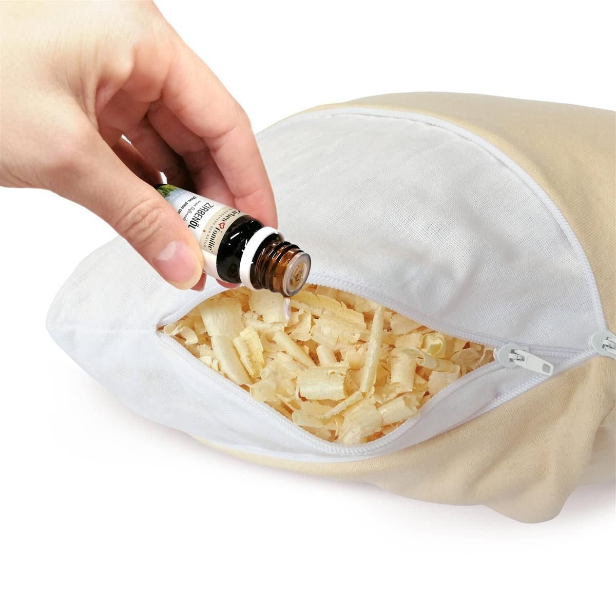 Beiges, herzförmiges Kissen mit Reißverschluss, geöffnet. Innen Holzspäne. Eine Hand gießt Öl aus einer Flasche.