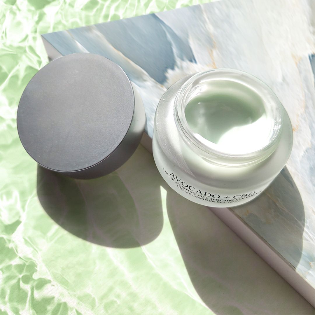 Augencreme-Tiegel mit geöffnetem Deckel. Aufschrift: Avocado + CBD, 8-Hour Moisture Fill Avocado Eye Cream, Crème Contour des Yeux. Hintergrund.