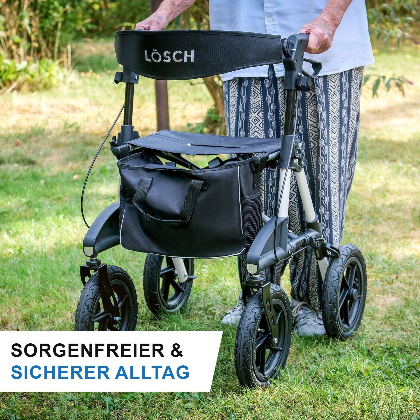 Person mit Rollator im Freien. Schwarzer Sitz und Tasche. Schwarze Räder, silberner Rahmen. Marke: Lösch.