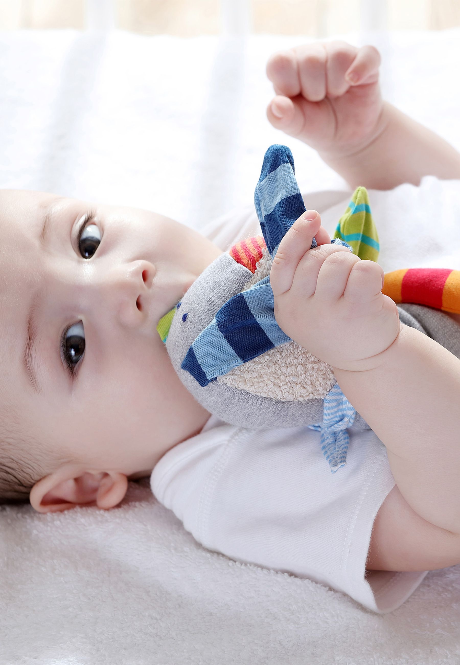 Baby liegt auf weißer Decke und hält Schnuffeltuch. Das Tuch hat bunte Details und ist im Mund des Babys.